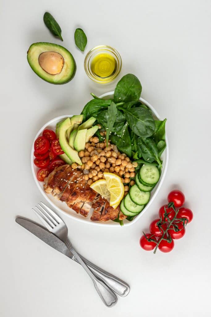 Bol cœur rempli de poulet grillé, avocat, épinards frais, pois chiches, concombres et tomates, avec huile d'olive et couverts.