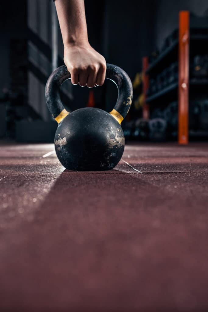 Prise puissante sur une kettlebell noire et jaune usée, posée sur le sol granuleux d'une salle de sport. Entraînement de force.