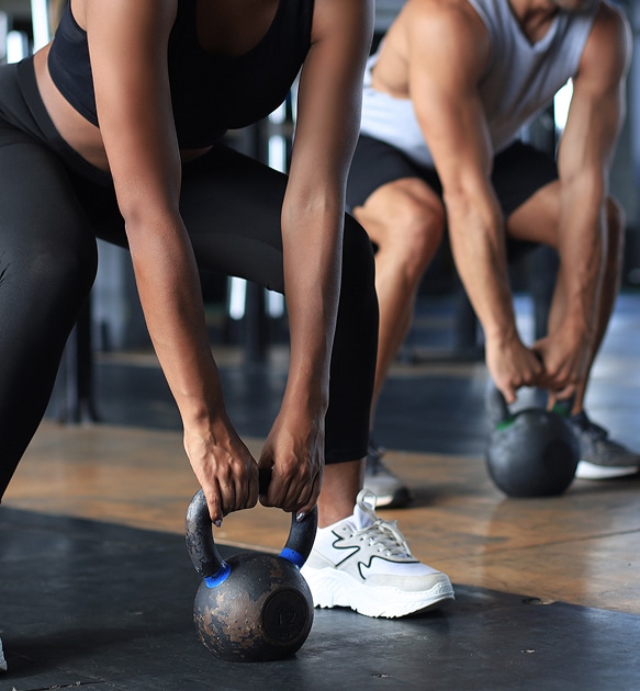 Kettlebell : entraînement intense pour le squat et le fitness. Femme en squat profond saisissant un kettlebell usé (noir/bleu) dans un gym, s'entraînant avec un homme en arrière-plan.