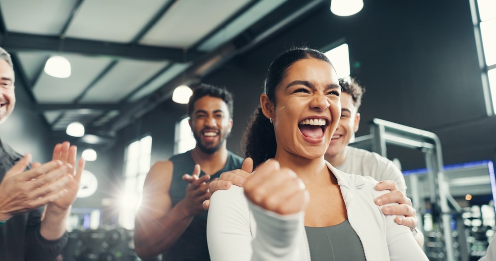 Succès fitness : Célébration joyeuse au gym en équipe. Femme joyeuse levant le poing, célébrant un succès fitness au gym, acclamée par son équipe souriante qui applaudit.